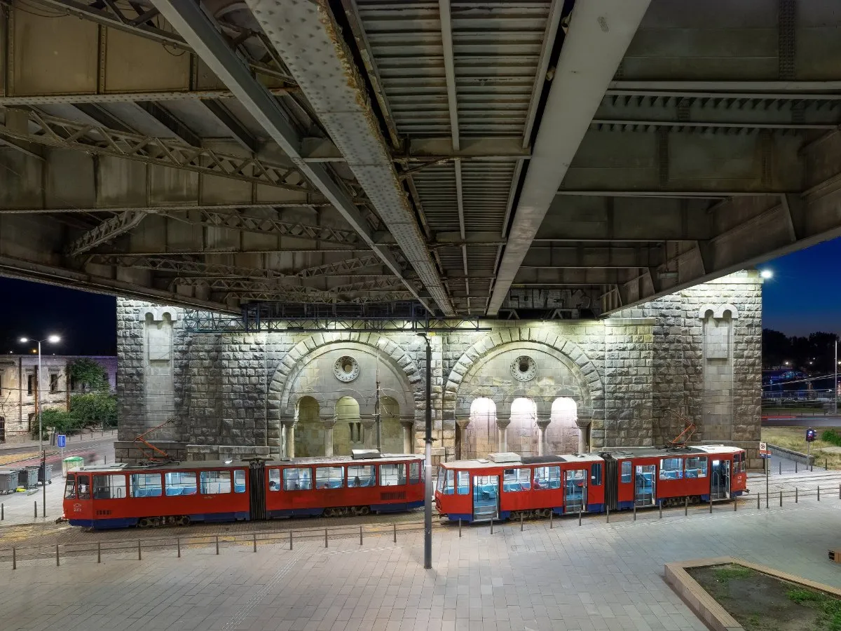 Two red and white trams wait at the stop under the steel girders of Branko's Bridge in Belgrade at dusk, in front of a lit stone arch.
