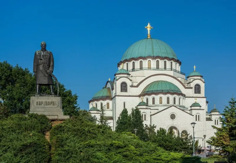 The white Church of Saint Sava with green copper domes rises behind the bronze statue of Karađorđe and the trees of the park, under a clear blue sky.
