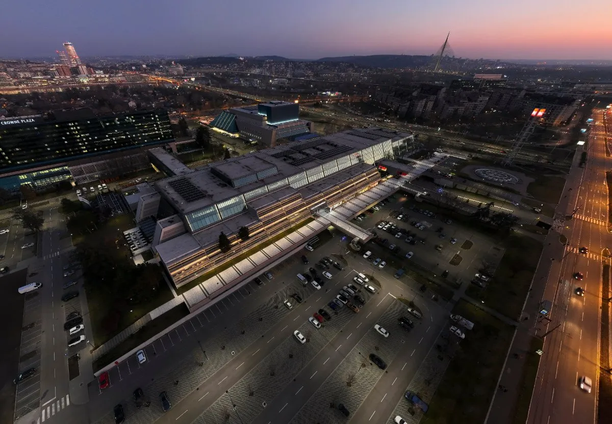 An aerial view at night of the long, lit Sava Centar congress building in New Belgrade, with car parks, avenues and city lights under a pink evening sky.