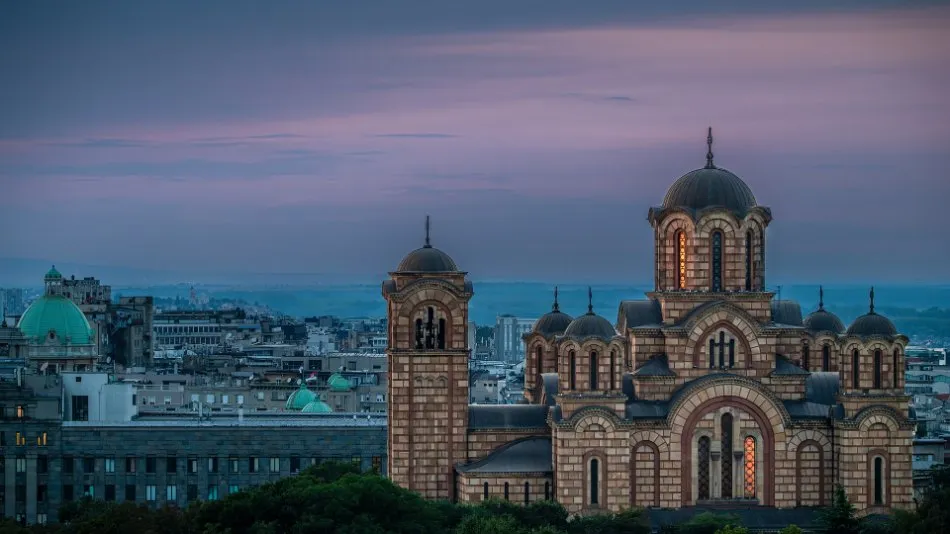 The red and yellow brick St. Mark's Church in Belgrade at dusk, with lit windows and grey domes above the roofs of the city under a pink sky.