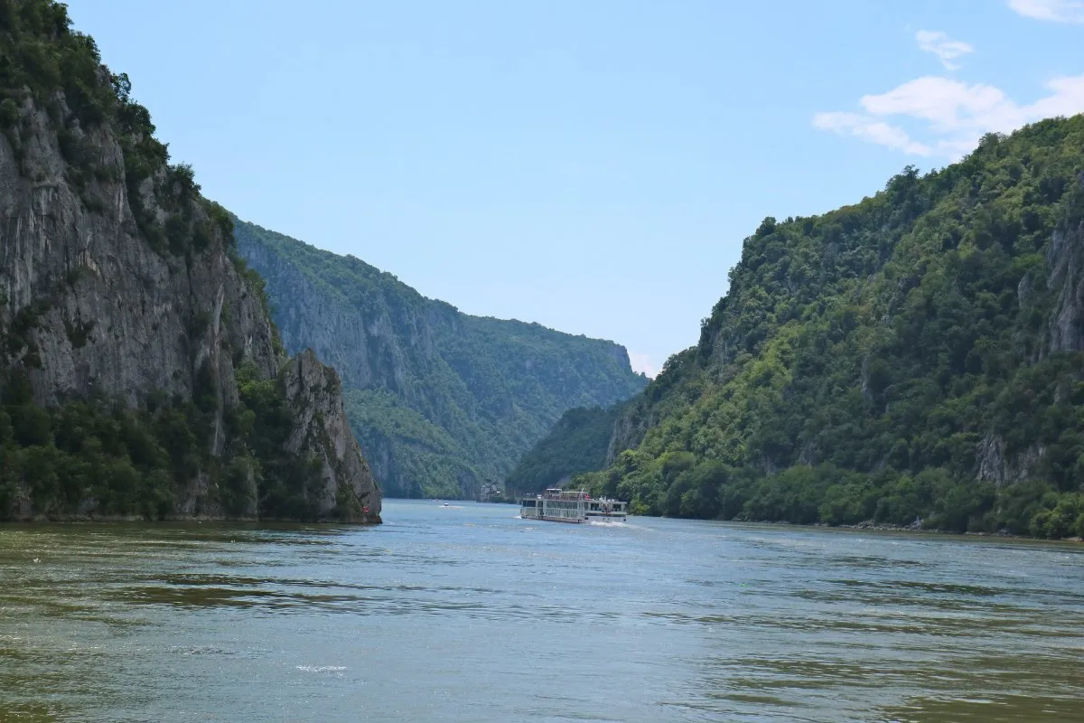 The Danube between steep wooded cliffs at the narrowest point of the Đerdap gorge, with a white river boat far off.