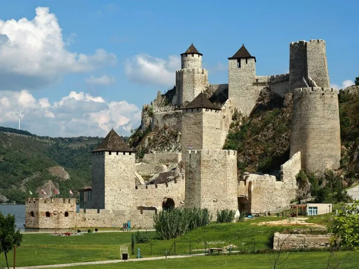 The stone towers and walls of the medieval Golubac Fortress climb a rocky hill at the entrance to the Đerdap gorge, with a green lawn in front and white clouds above.
