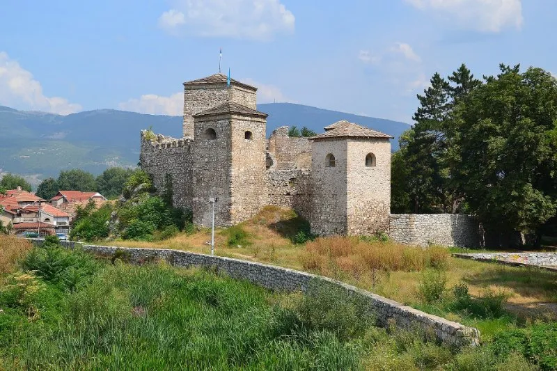 The stone towers of the Pirot Fortress with their pyramid roofs above green grass, with houses and wooded mountains behind.