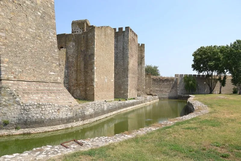 The tall stone towers and walls of the 15th-century Smederevo Fortress above a green moat, under a clear blue sky.