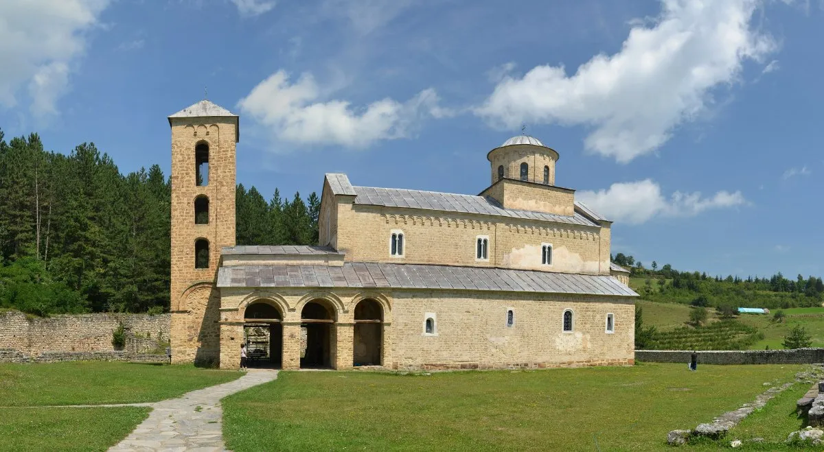 The 13th-century church of the Sopoćani Monastery with its bell tower and arched porch on a green lawn, with pine forest behind and white clouds in a blue sky.