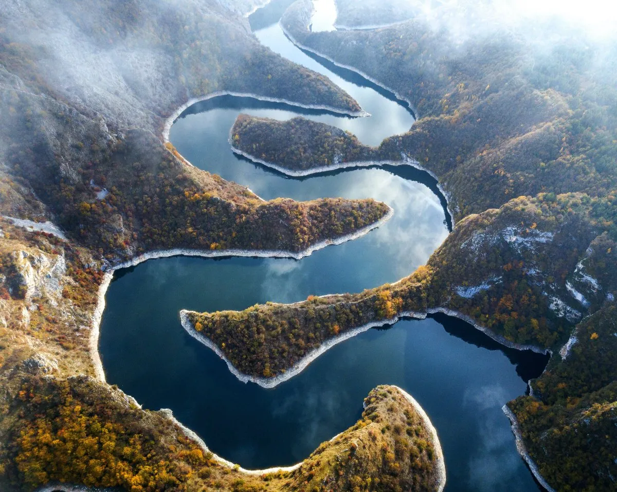 An aerial view of the dark blue Uvac river turning in tight loops through a canyon of autumn forest and pale limestone, with mist on the slopes.
