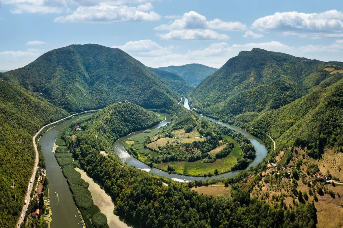 An aerial view of the West Morava making a wide loop around a green meadow between the forested mountains of the Ovčar-Kablar gorge.